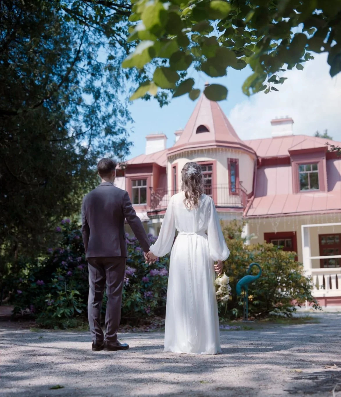 A couple, dressed in wedding attire, holding hands and walking away from the camera towards a large pink house with a turret. The woman is in a white wedding dress holding a bouquet, and the man in a gray suit. The scene is surrounded by greenery and