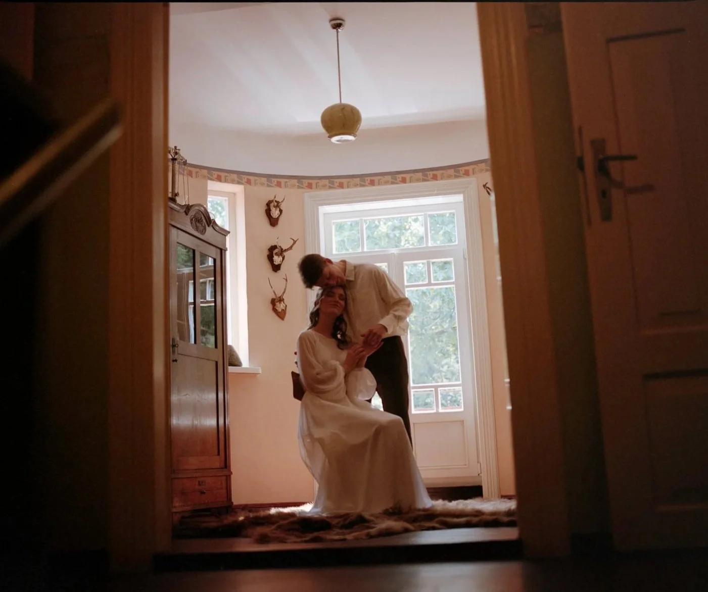 A couple in vintage clothing inside a room, with one sitting on a chair and the other standing, looking at each other, framed by a doorway.