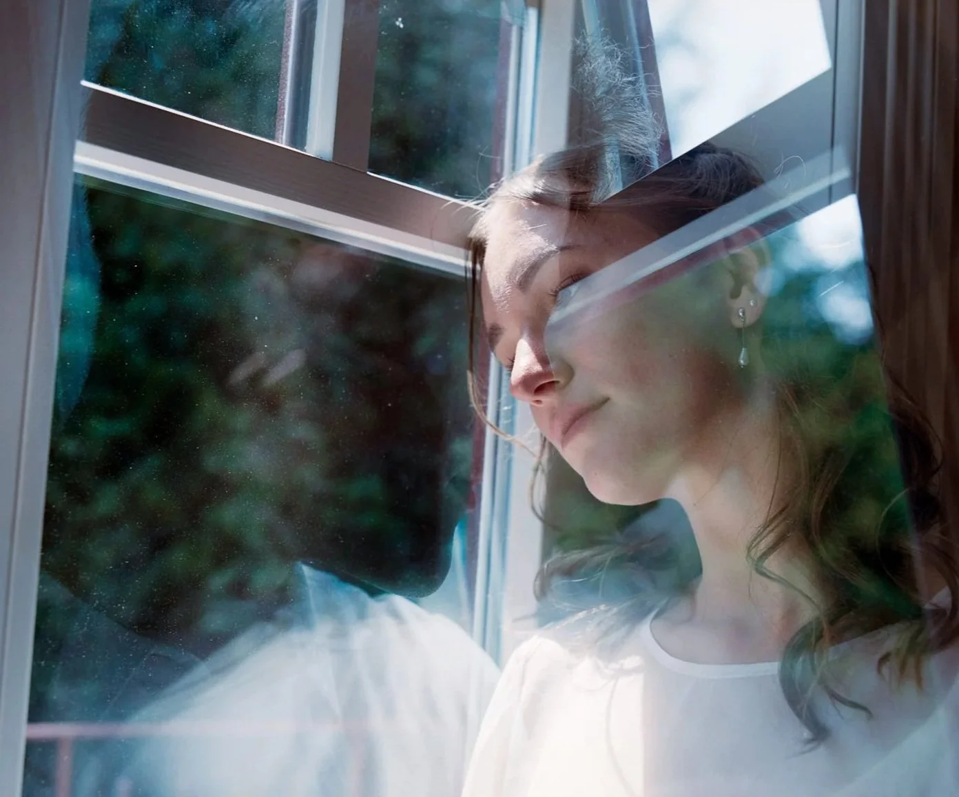A young woman with light skin and brown hair, wearing earrings, is looking out of a window with a smile. There is a reflection of a man with dark skin and curly hair on the window glass, and outside, there are trees.