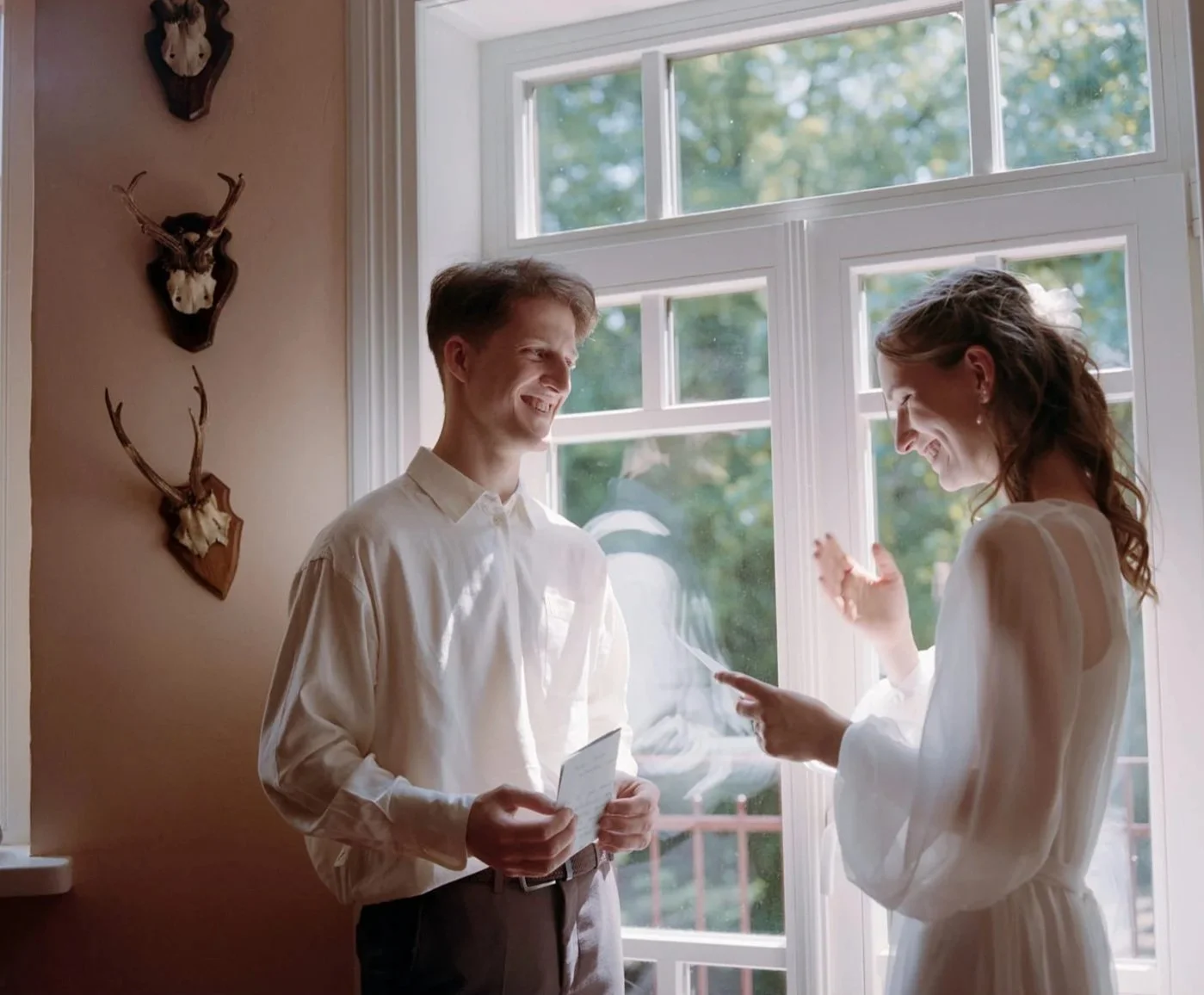 A man and woman sharing a joyful moment indoors by a large window. The man is holding a letter, and the woman appears to be reading or showing something from a piece of paper. The room has deer head wall mounts as decor.