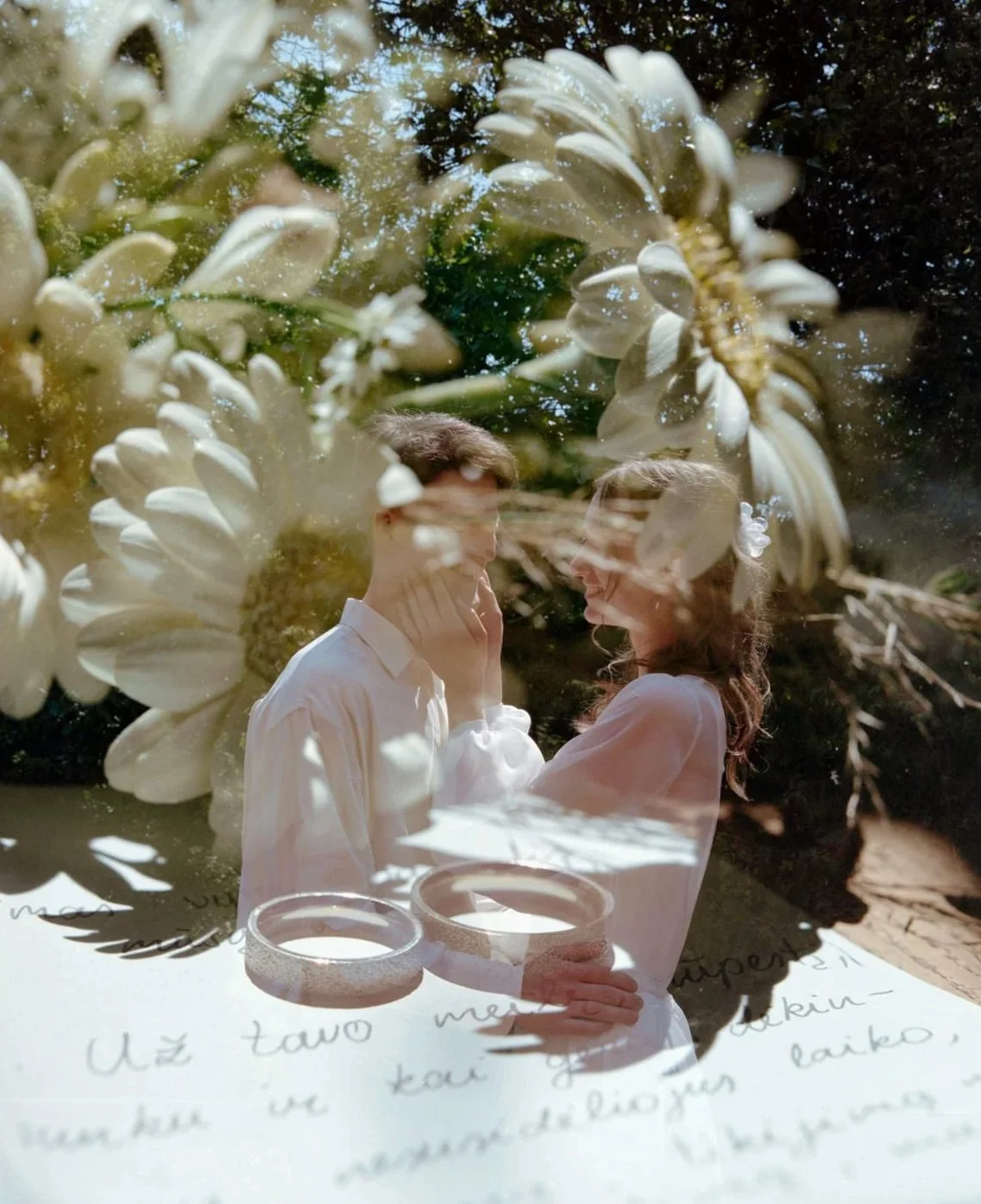 A double exposure photograph of a young couple smiling at each other, overlaid with white flowers, greenery, and handwritten text.