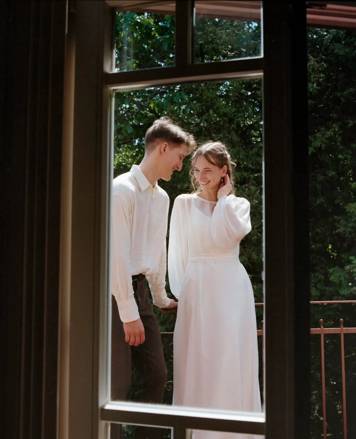 A couple dressed in vintage white clothing standing outdoors on a balcony, seen through a window.