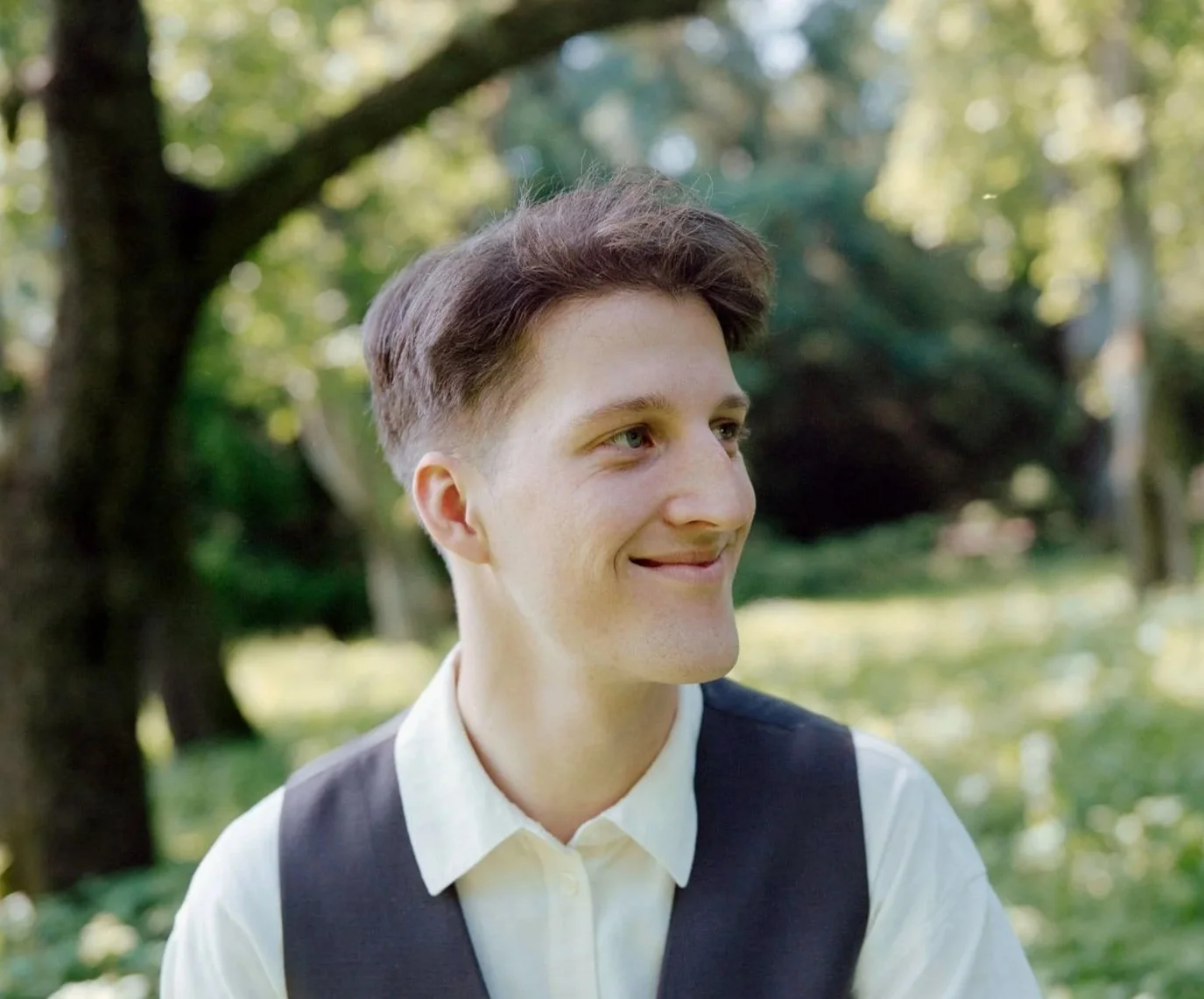 Young man with brown hair and light skin smiling outdoors in a park with trees and greenery in the background.