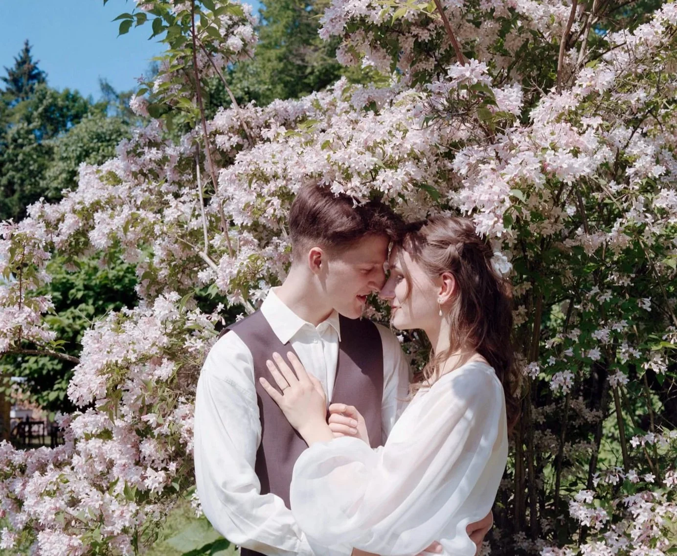 A young couple standing close together with foreheads touching, surrounded by pink cherry blossoms in a garden.