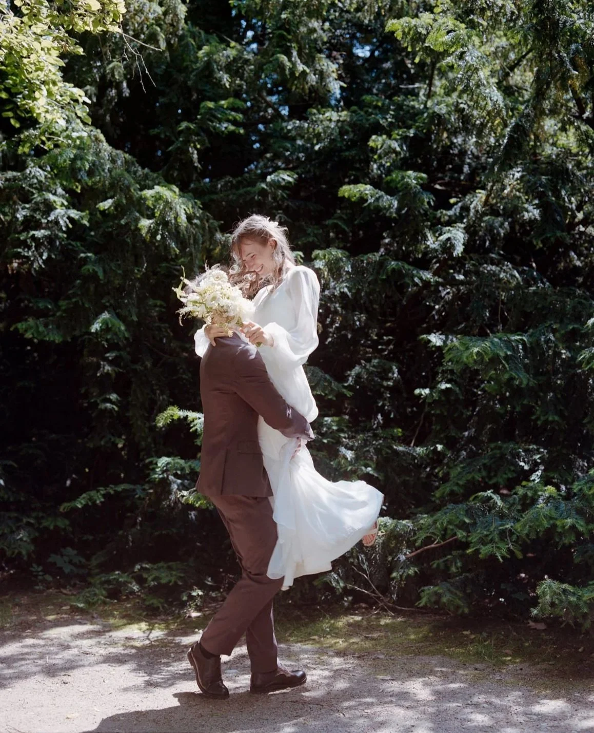 A couple dancing outdoors surrounded by green trees, with the woman in a white dress holding a bouquet of flowers, lifted by the man in a dark suit.