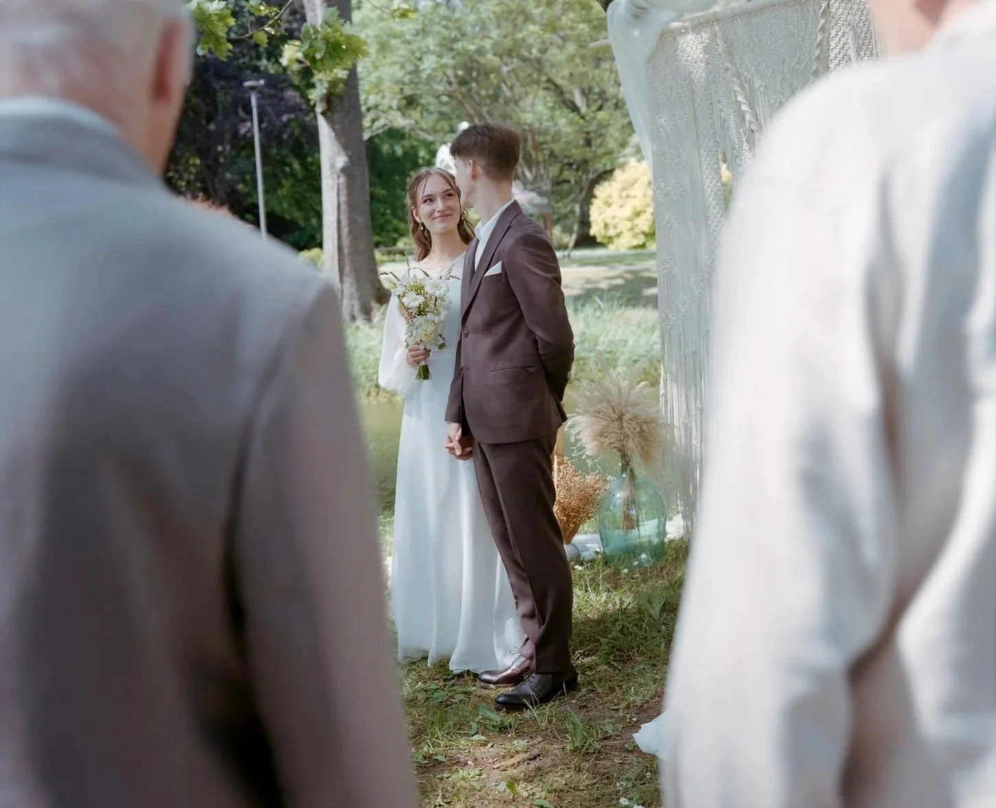 A couple getting married outdoors, with the bride holding a bouquet of white flowers and the groom wearing a brown suit. They are standing in front of a decorative backdrop, surrounded by greenery, with two people in the foreground observing the cere