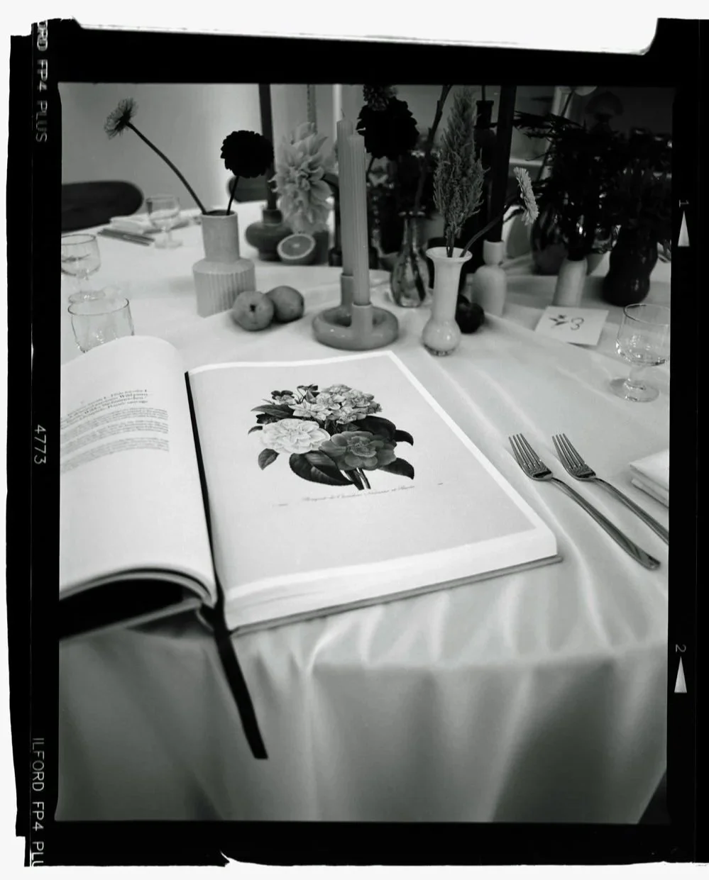 A table set for a meal with a floral open book, vases with flowers, apples, glasses, and cutlery, decorated in a minimalist style.