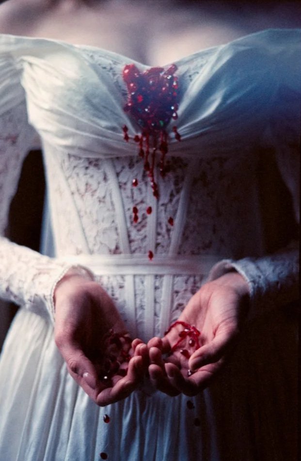 A person wearing a white wedding dress with blood on the bust and hands, holding red beads or jewels.