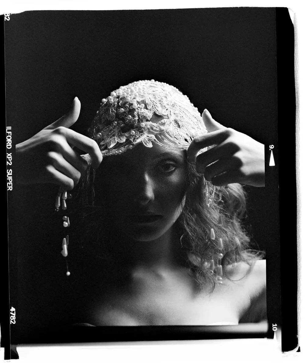 Black and white photograph of a woman wearing an ornate headpiece with lace and pearls, touching it with both hands, with dramatic lighting highlighting her face and upper body against a dark background.