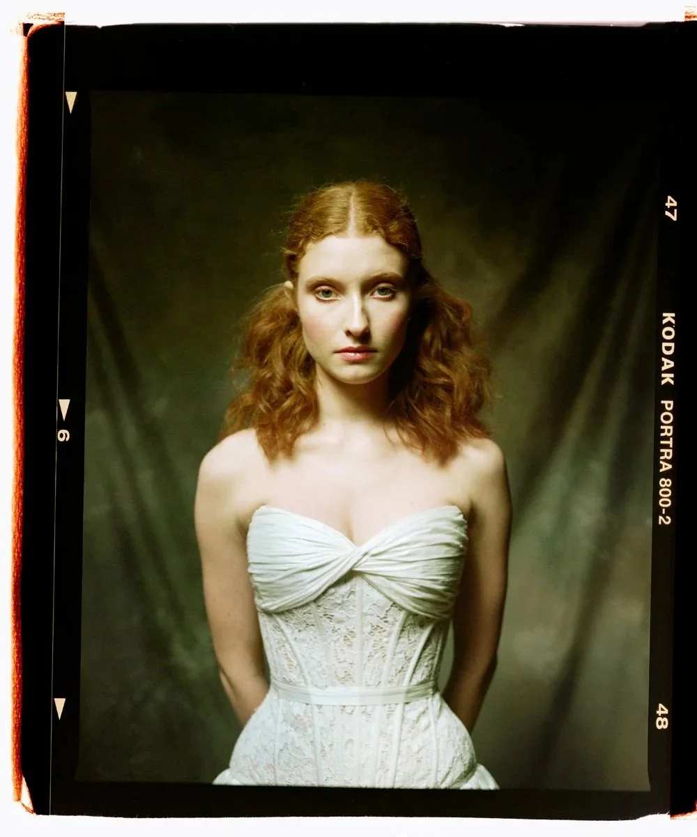 A portrait of a young woman with red hair wearing a white strapless dress with lace detailing, standing against a dark background.