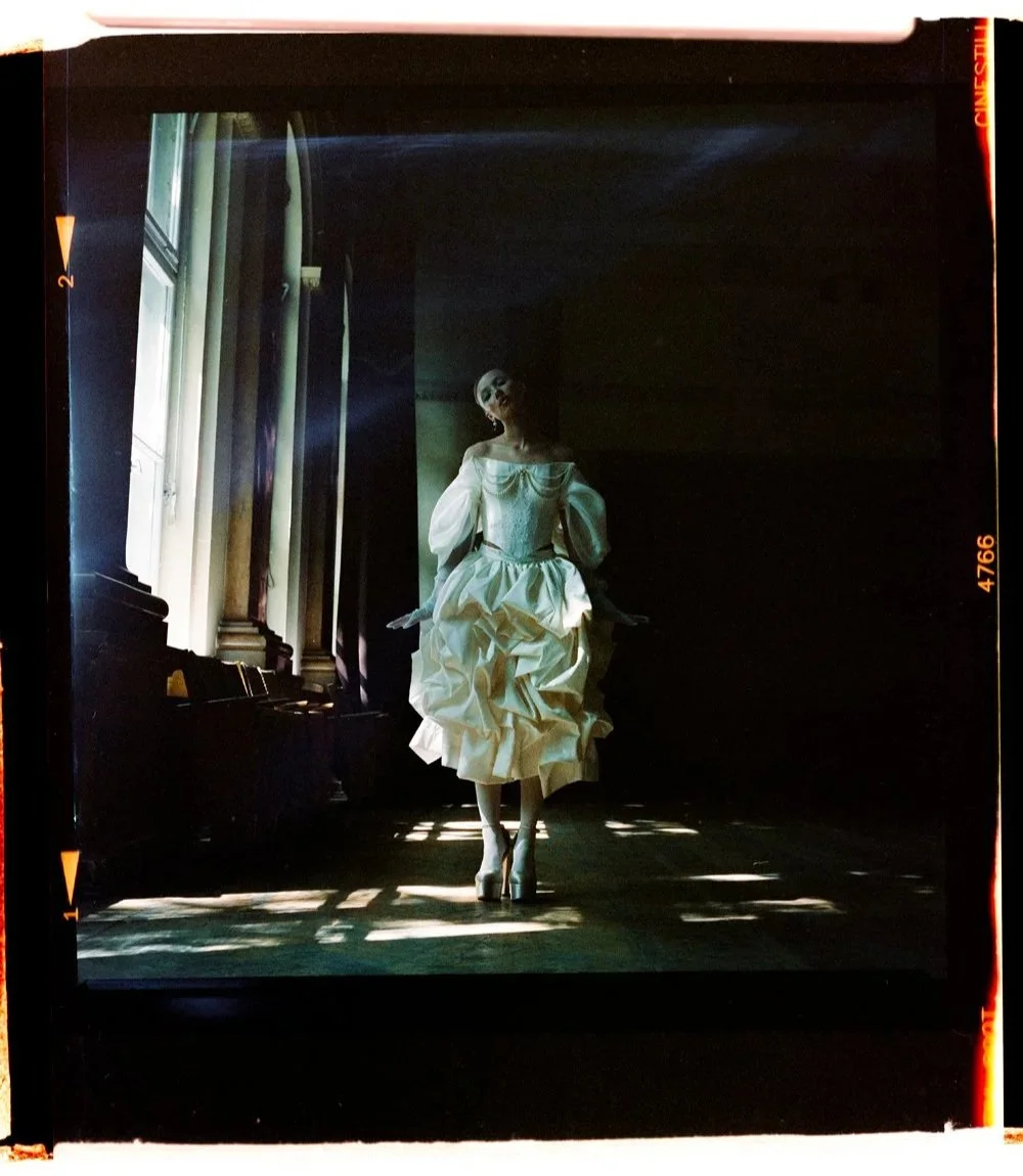 A woman in a vintage white dress with puffed sleeves and ruffles, standing in a dimly lit room near a window with sunlight streaming in.