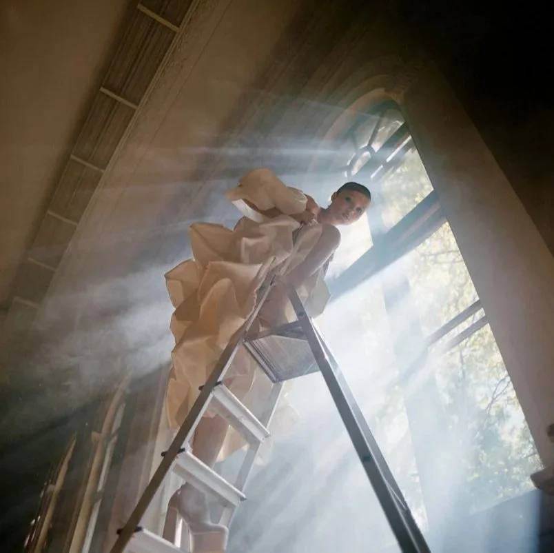 A woman in a light-colored dress standing on a ladder in front of a large window with sunlight streaming in, creating beams of light across the room.