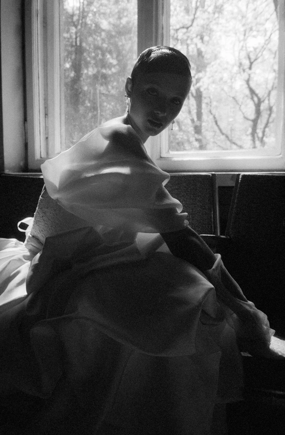 A black-and-white photo of a woman with short hair sitting on a bed, looking towards the camera. There is a window behind her, showing trees outside.