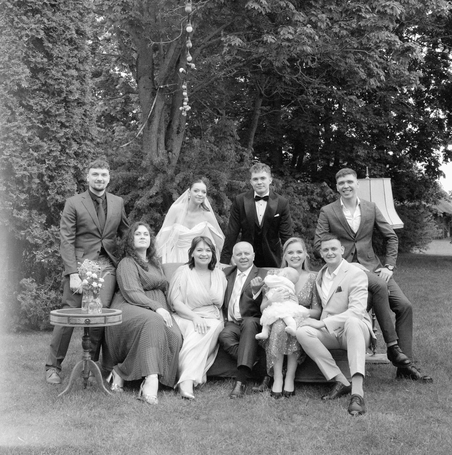 A large black-and-white wedding portrait of a family, with a group of ten people and a baby, outdoors near trees and a garden. Three women and seven men, one of the women is wearing a wedding dress and veil, and some individuals are holding bouquets 