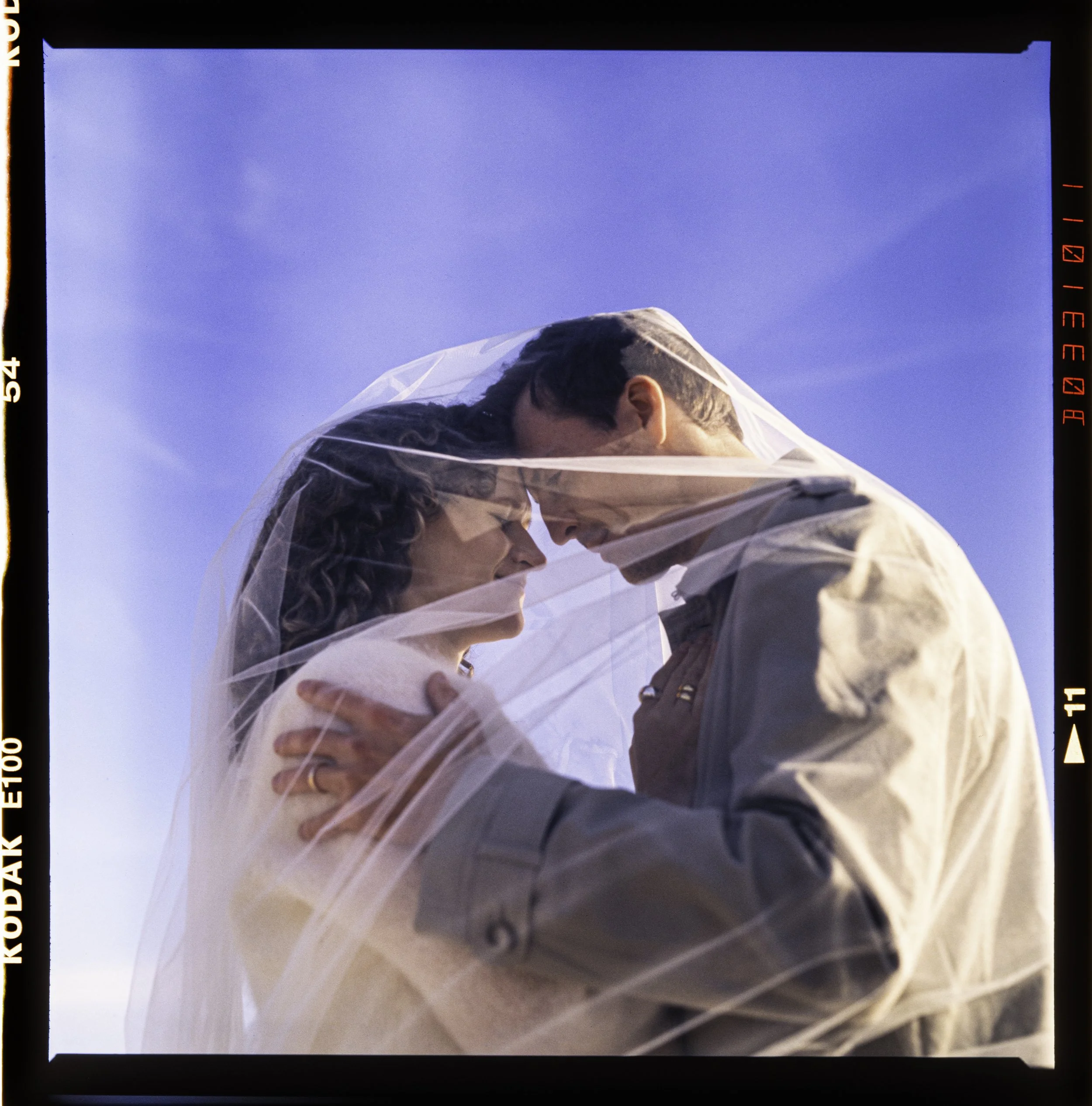 A bride and groom embrace under a sheer veil, touching foreheads and holding hands, against a clear blue sky.