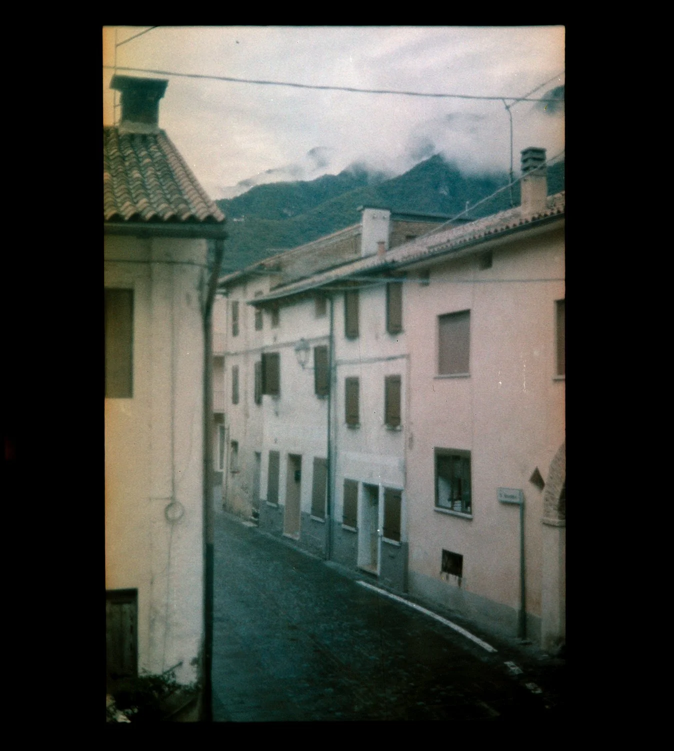 A narrow street in a European town with pastel-colored buildings and green shutters, with mountains and cloudy sky in the background.