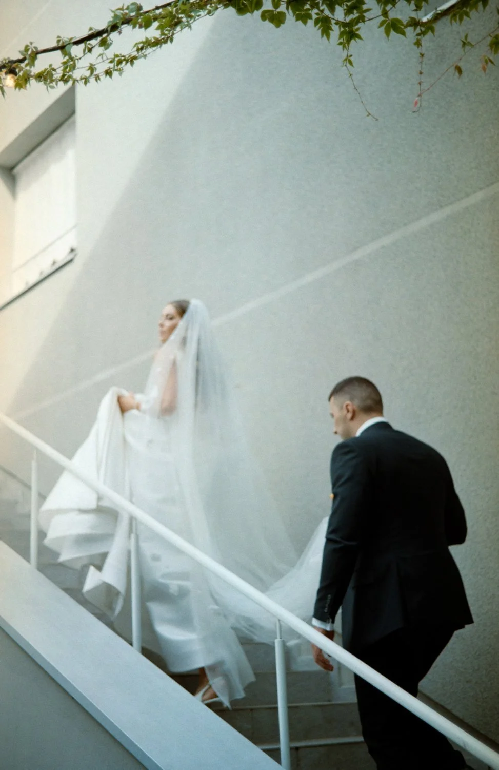 A bride in a white wedding dress and veil walking up a staircase, with a groom in a black suit following behind her.
