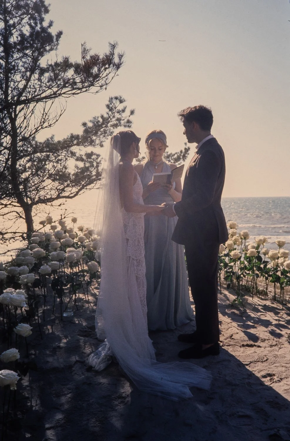 A couple getting married outdoors by the beach at sunset, standing with an officiant, holding hands, with white flowers and trees in the background.
