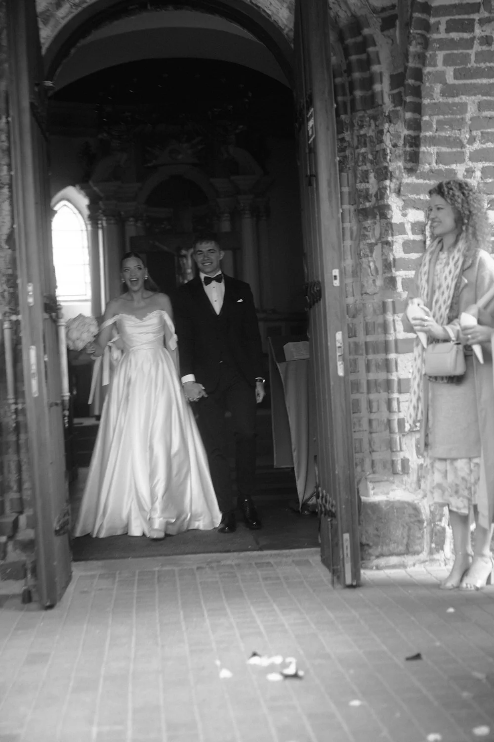 A wedding couple, a bride in a white dress and a groom in a tuxedo, walking through a church door, smiling, with guests on the side.