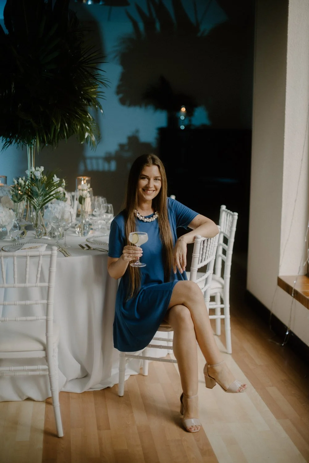 A woman in a blue dress and heels sitting at a decorated dinner table during an evening event, holding a cocktail and smiling.