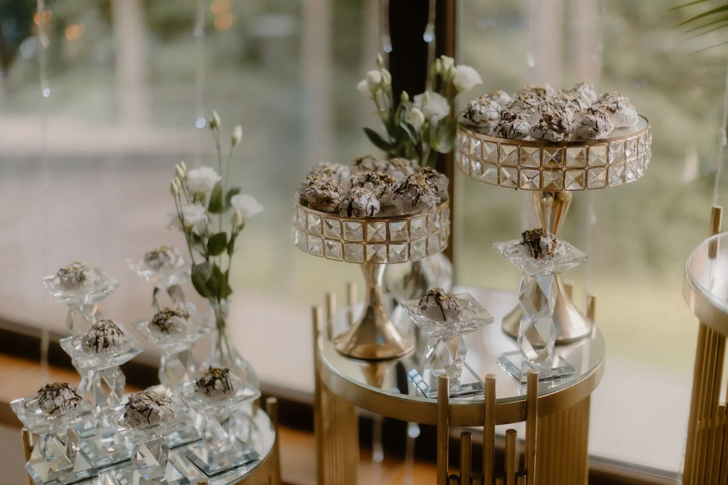 Elegant dessert display with cookies on gold and glass stands, decorated with white flowers, set near large windows with a view of greenery outside.