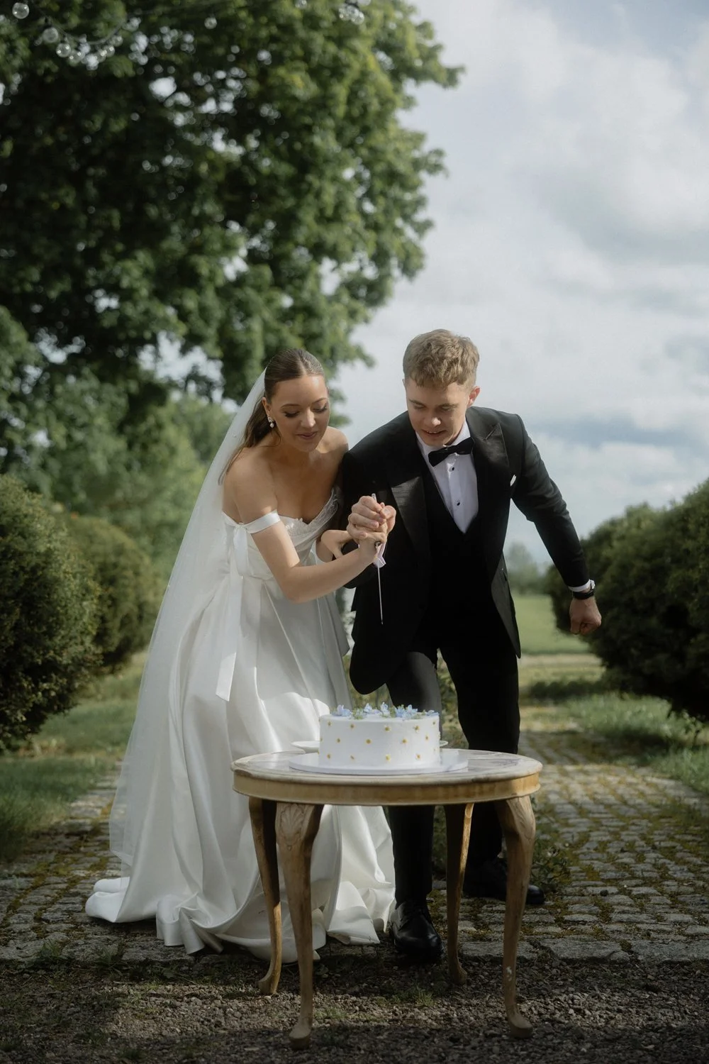 A newlywed couple in wedding attire, the bride in a white dress and veil, the groom in a black tuxedo, cutting a wedding cake outdoors on a small wooden table under a cloudy sky with trees in the background.