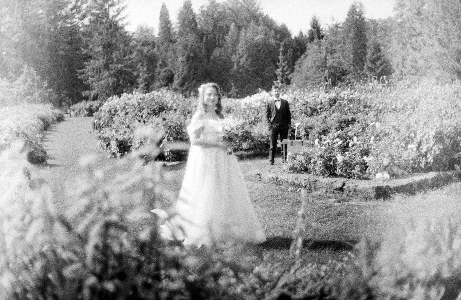 Black and white photo of a woman in a wedding dress and a man in a suit standing in a garden with flowers and trees.