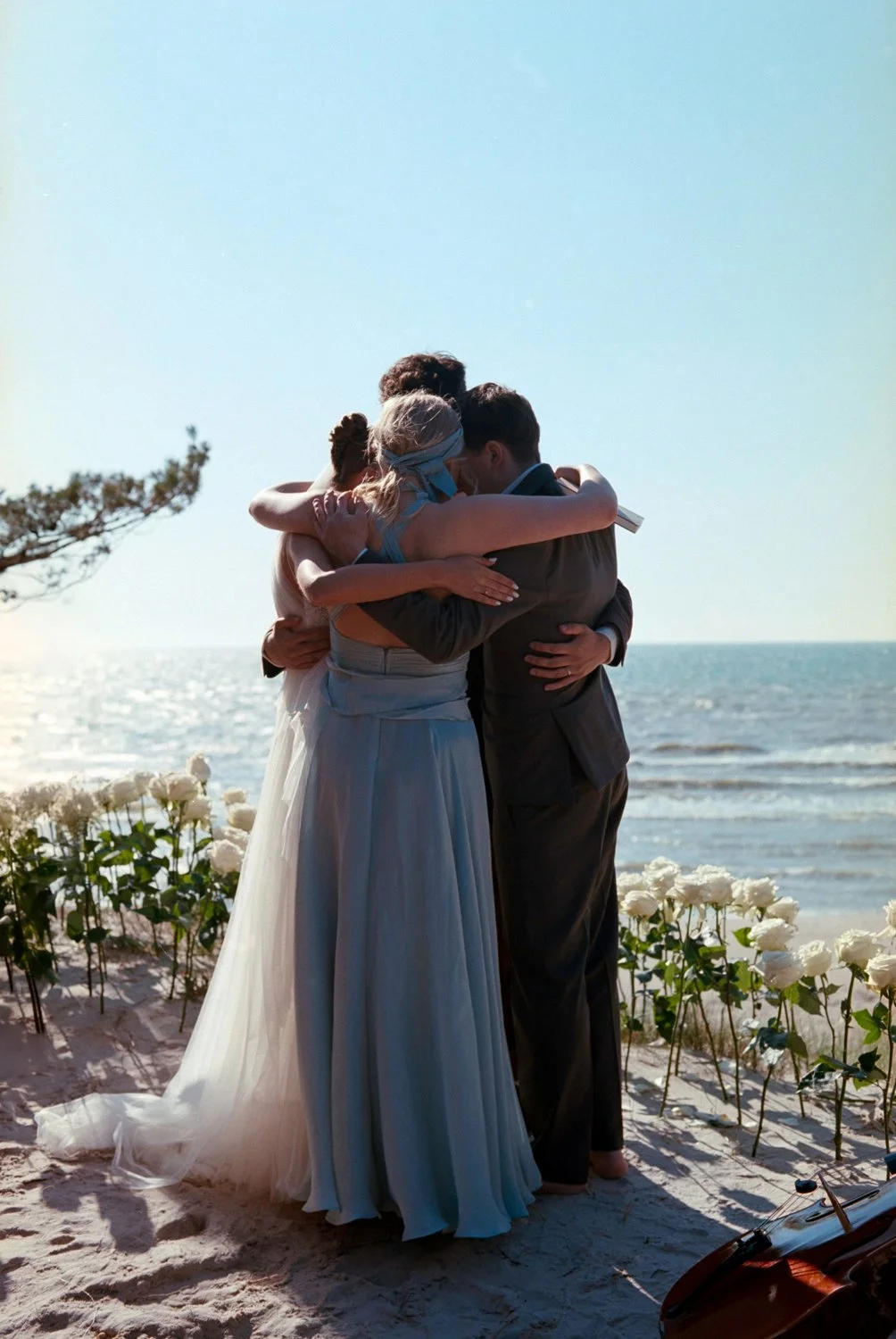 A group hug among a bride, groom, and two other women on a beach during sunset, with white flowers and a violin nearby.