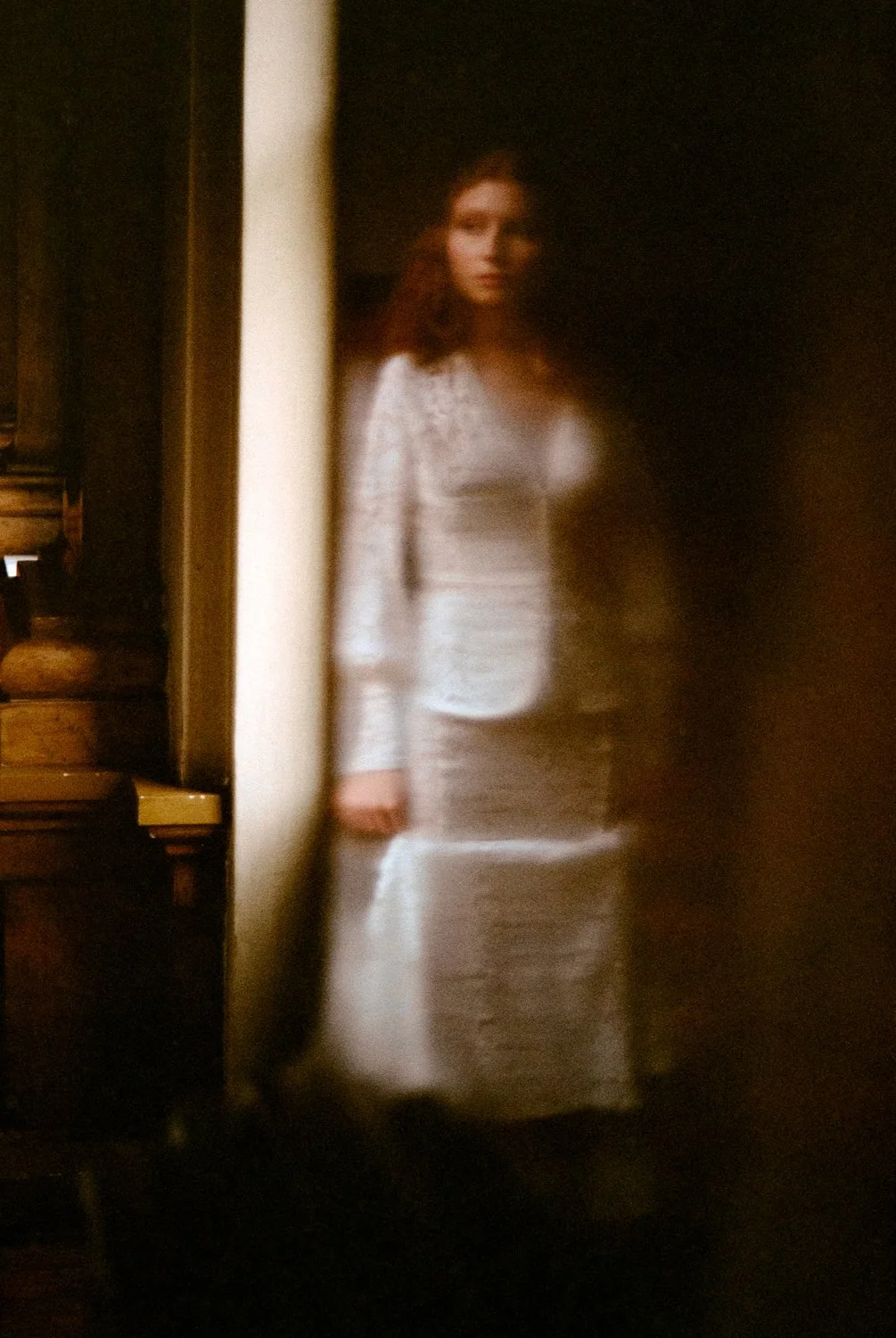 A woman with red hair standing in front of a mirror, wearing a white lace dress, with a blurred and dark background.