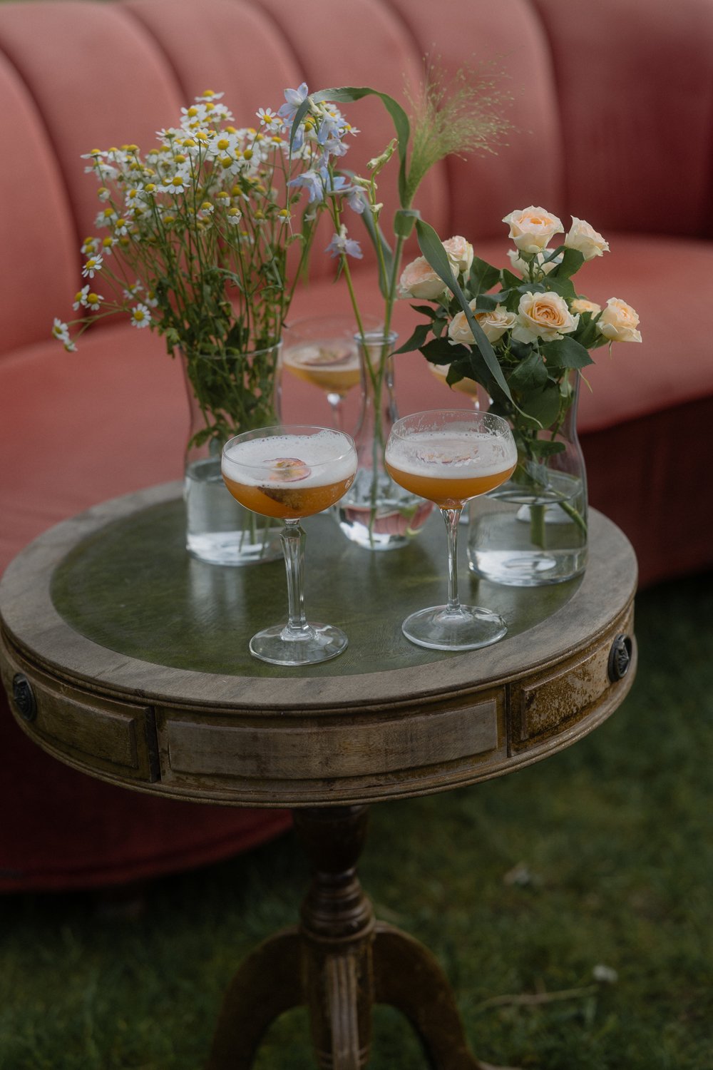 Three cocktail glasses with foam and garnishes on a small round wooden table with vases of wildflowers, set against a pink cushioned bench.