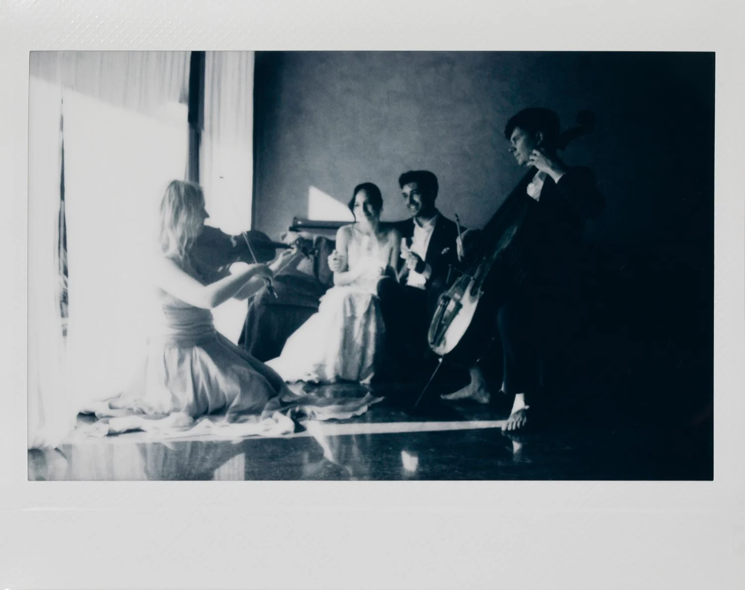 A black and white photo of four people in a room, with two playing a violin and guitar, and the other two sitting on the floor.