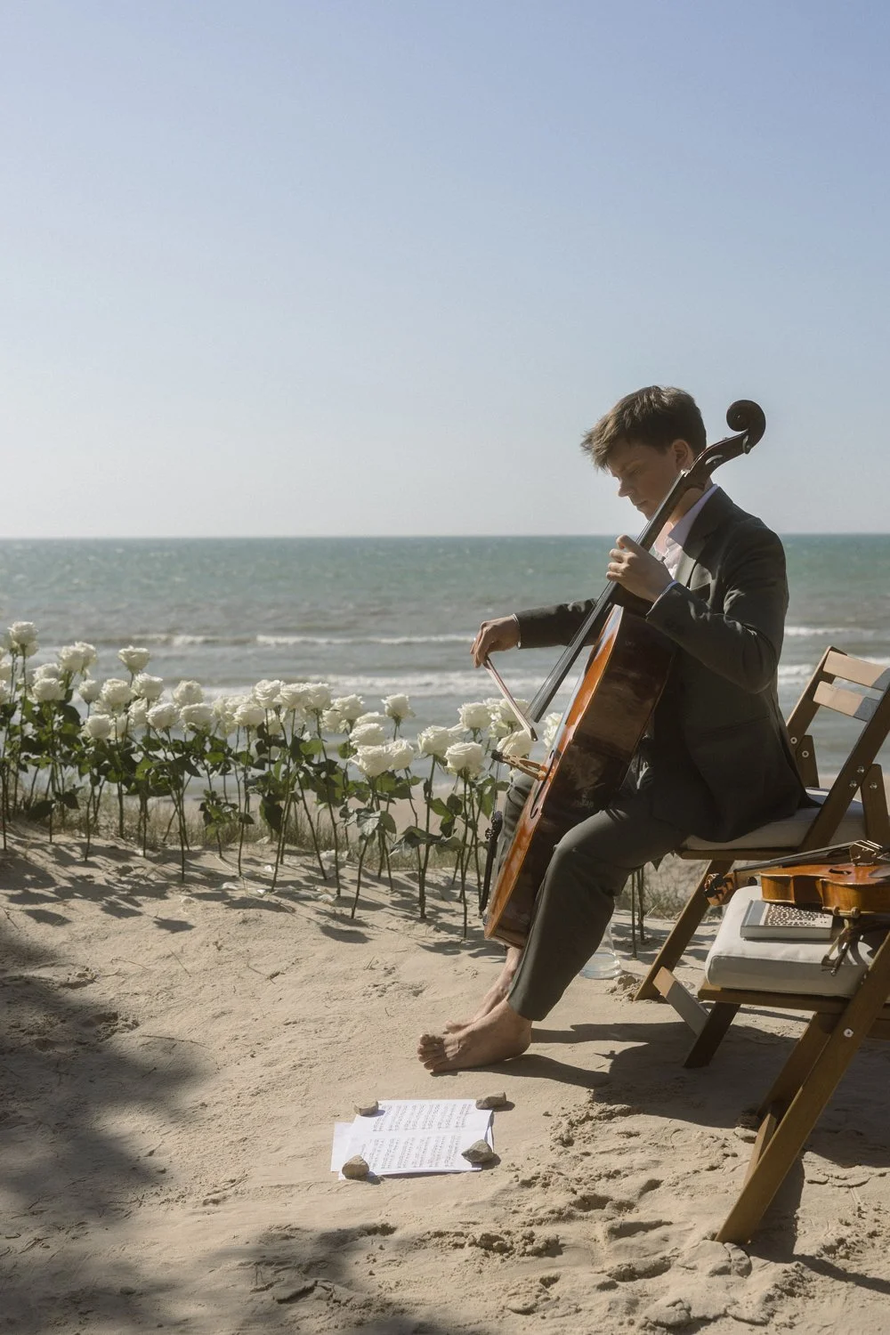 A young man dressed in a tuxedo playing a cello on a sandy beach near the ocean, with white flowers and a chair nearby.