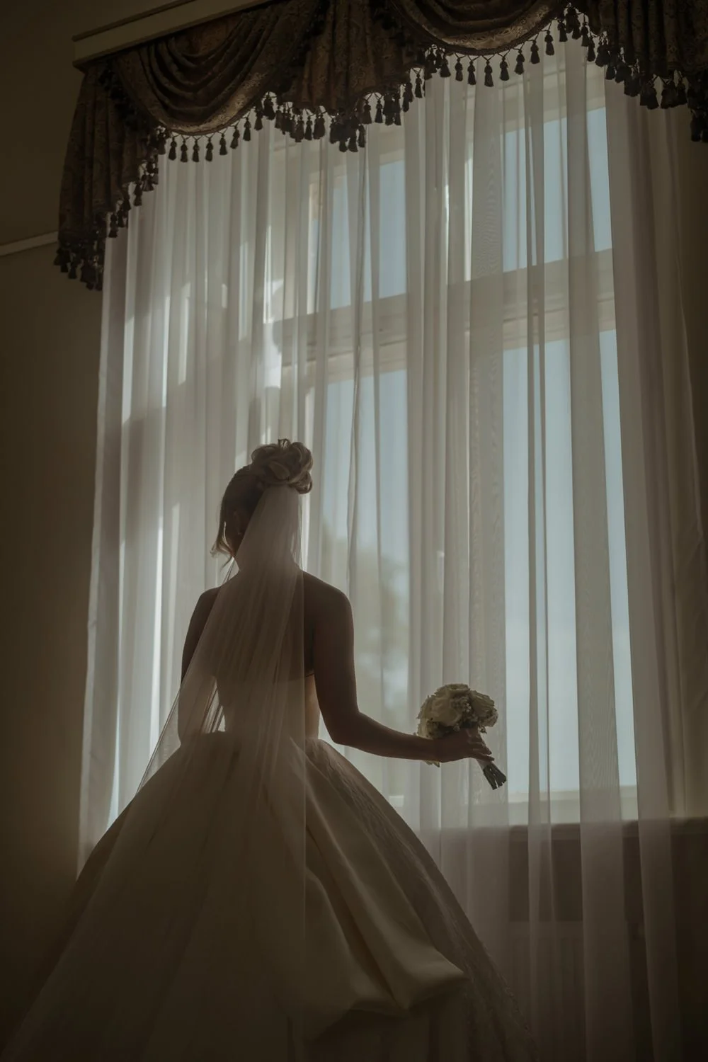 A bride in a wedding dress standing indoors near a large window with sheer curtains, holding a bouquet of flowers, with her face obscured by a veil.