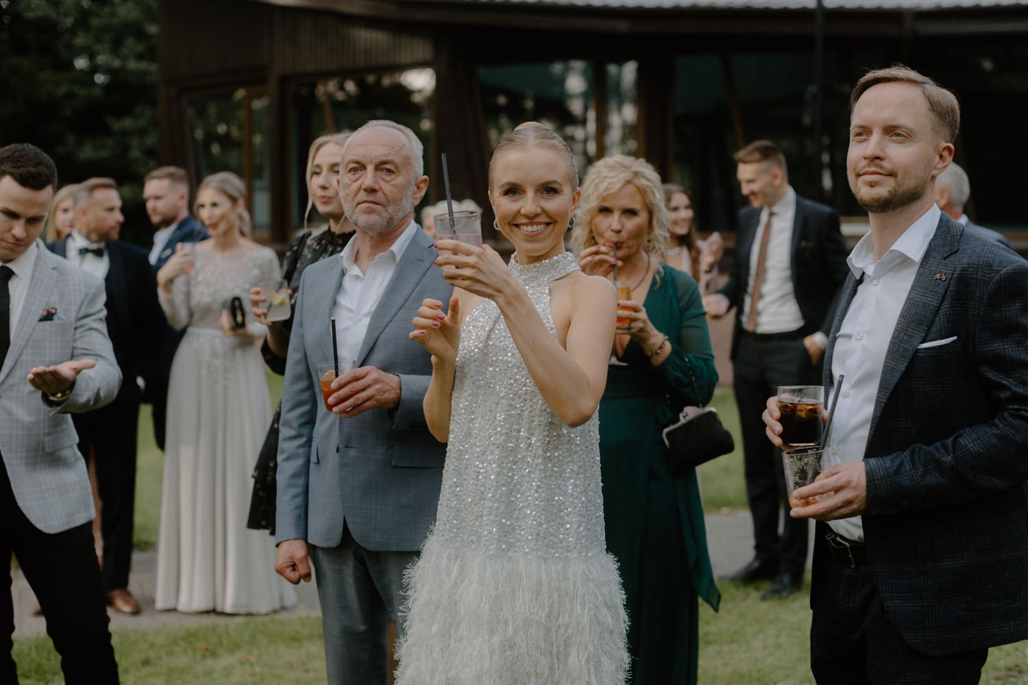 Wedding reception with a group of well-dressed people outdoors, including a woman in a sparkly white wedding gown smiling and holding a drink, surrounded by friends and family in formal attire.