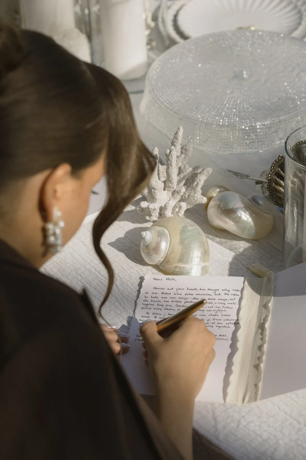 A woman in elegant earrings writes a handwritten note at a table decorated with seashells, coral, and a glass dish.
