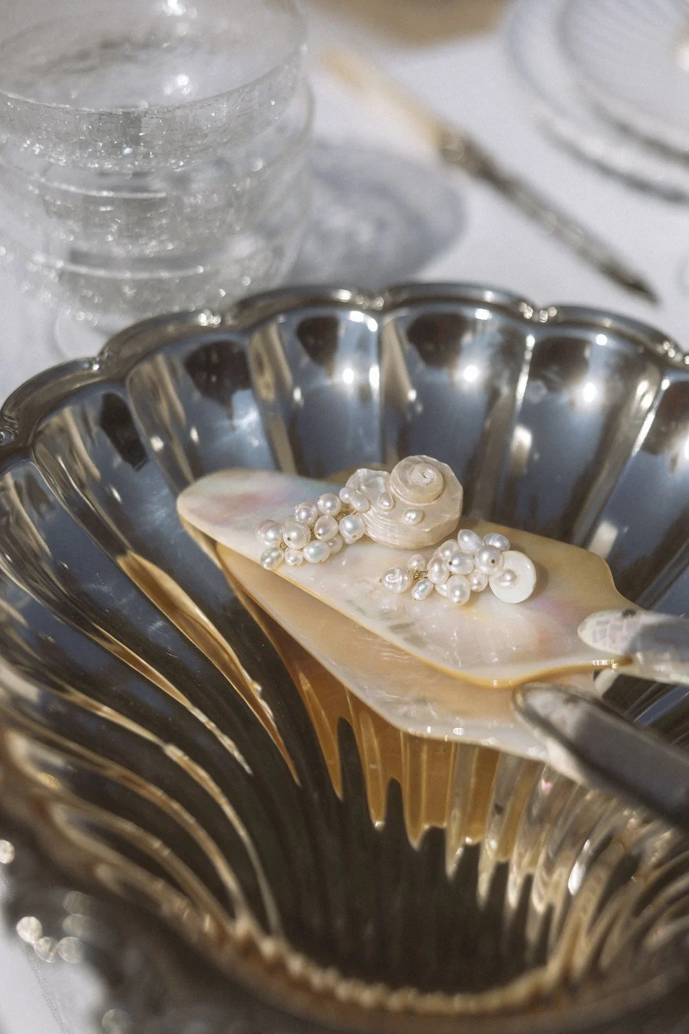 A silver serving dish with pearl jewelry and a shell-shaped ornament on a pearlescent slide resting on it. In the background, a stack of clear glass cups and in the top right, a plate and a knife.