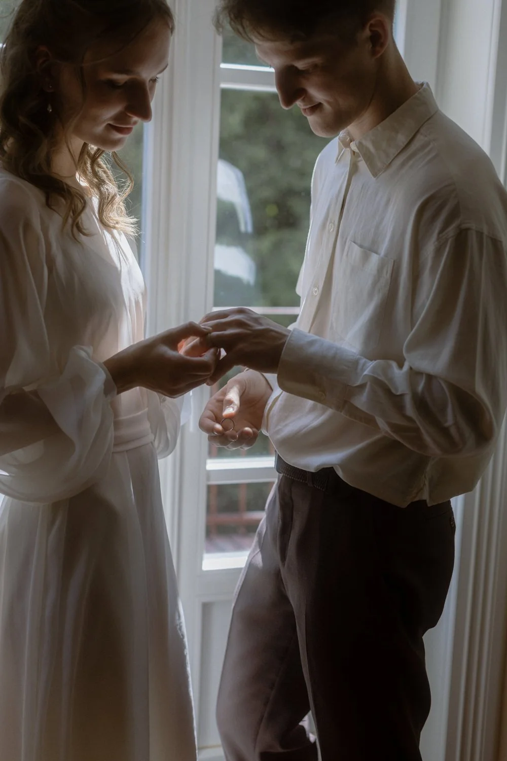 A couple is standing near a window, with the woman placing a ring on the man's finger during a private, intimate moment.