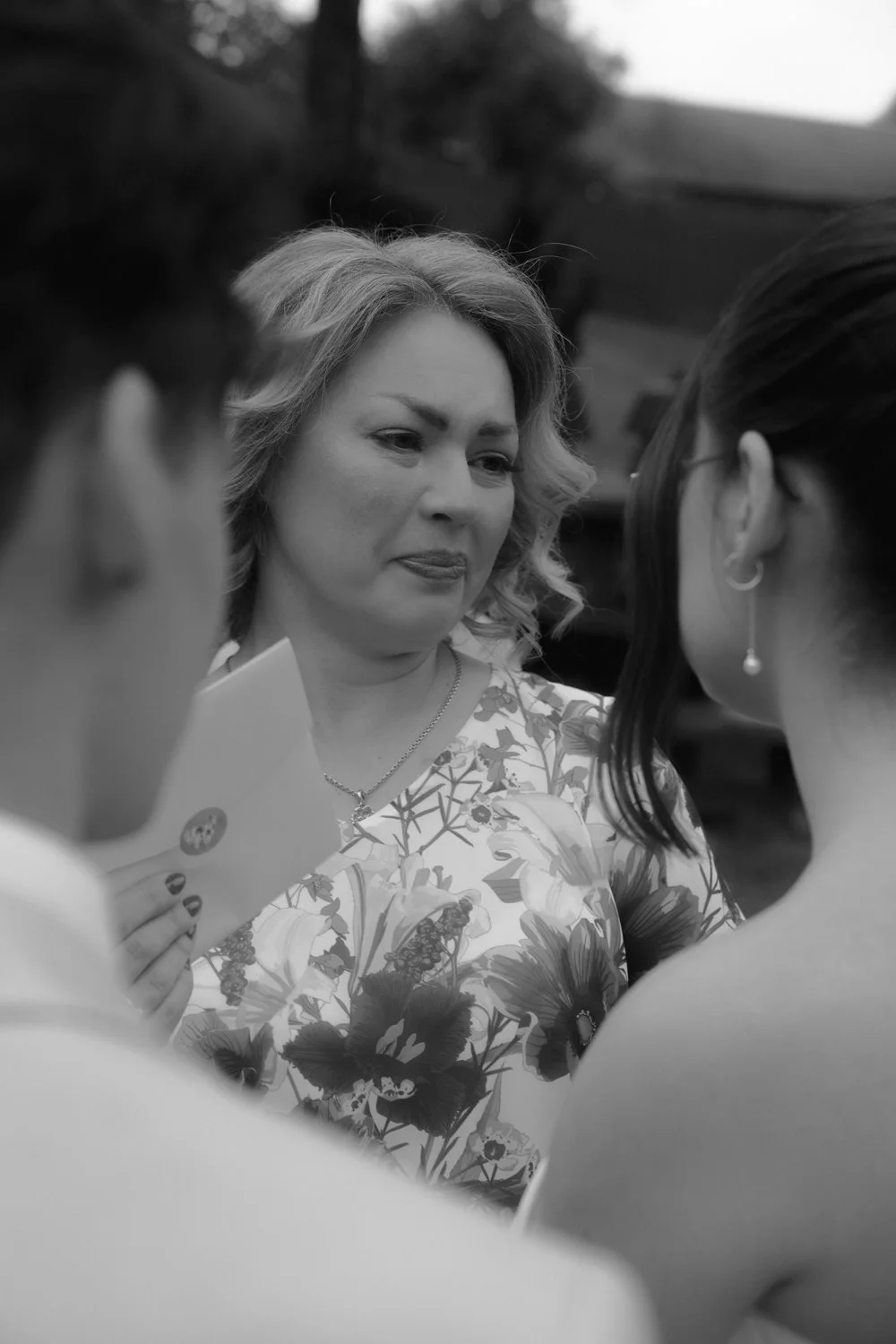 Black and white photo of a woman with curly hair wearing a floral blouse, holding a card, surrounded by two women, one with glasses and earrings, at an outdoor event.