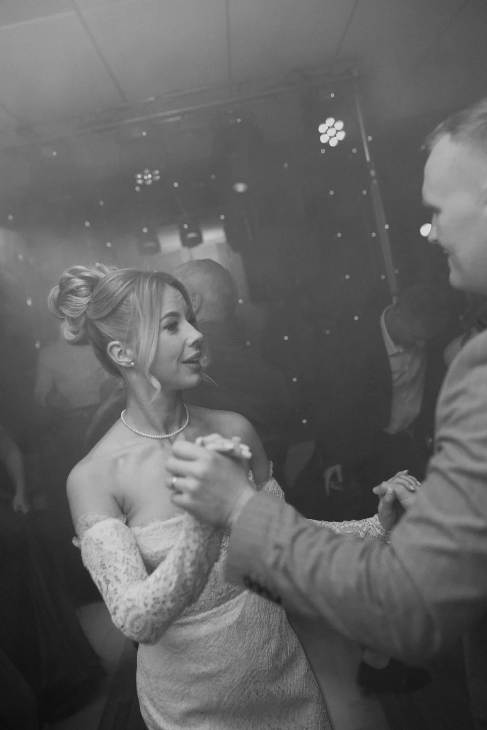 A bride in a strapless lace wedding dress and pearl necklace dancing with a groom in a suit, holding hands, in a dimly lit wedding reception.