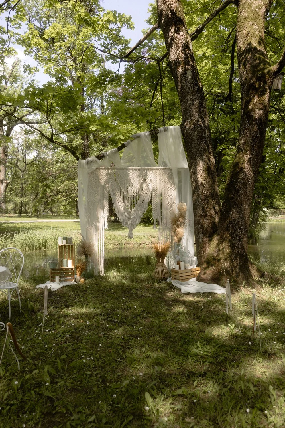 Outdoor wedding or event setup in a wooded area with a decorated arch draped with white fabric and macrame. Surrounding the arch are candles, dried flowers, and wooden crates, with a white metal chair nearby and a pond in the background.