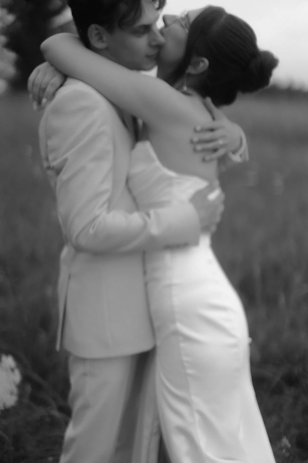 A black-and-white photo of a couple embracing outdoors, with the woman kissing the man on the lips, both dressed in light-colored clothing.
