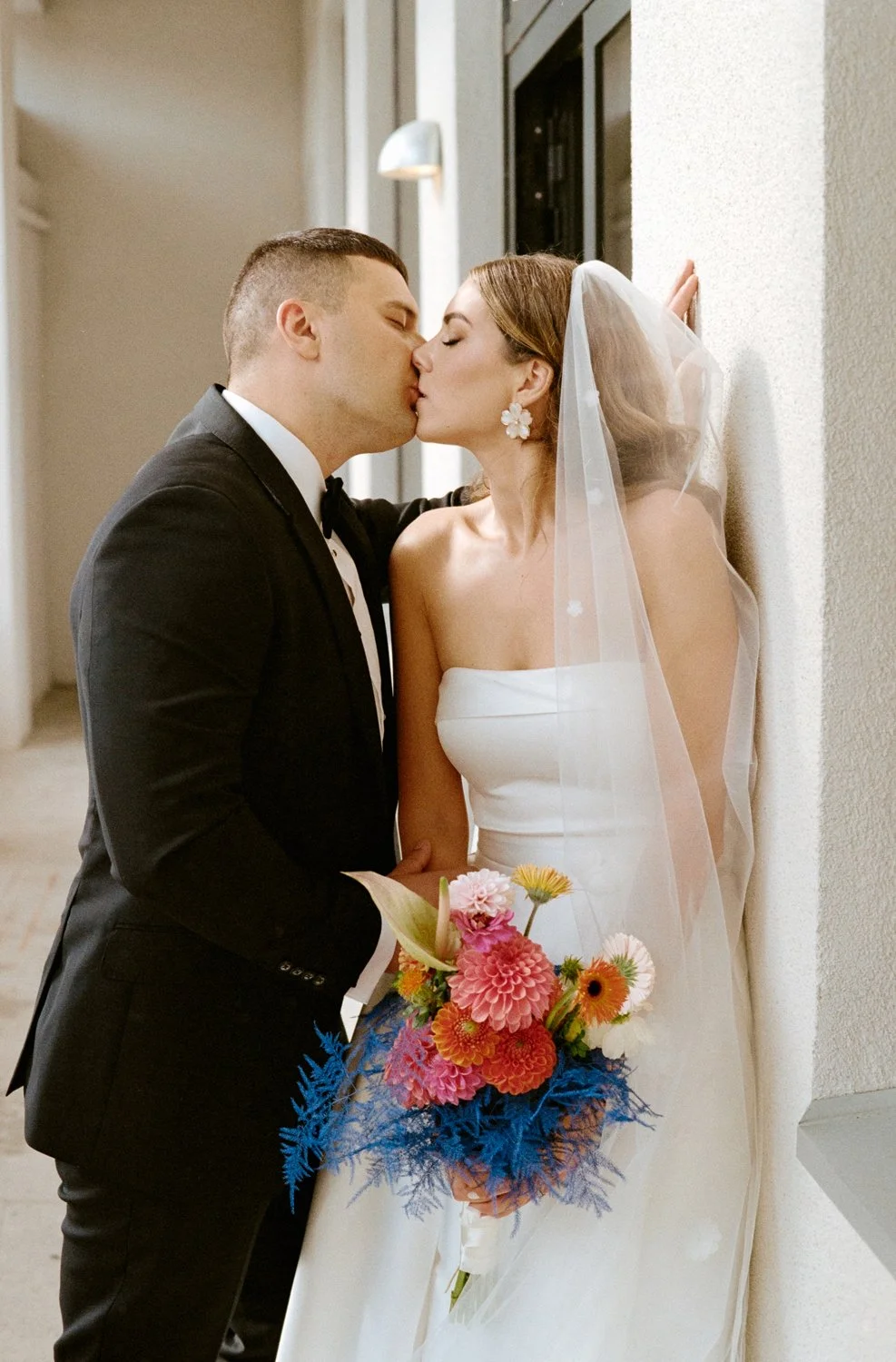 A bride and groom sharing a kiss in a hallway. The bride is holding a colorful bouquet and wearing a white strapless wedding gown with a veil. The groom is in a black suit with a white shirt and black bow tie.