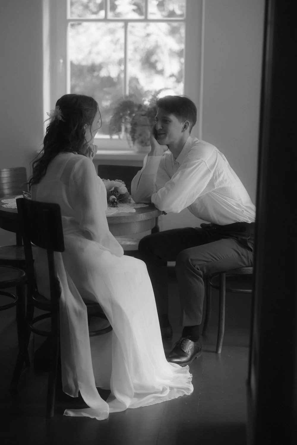 A black and white photo of a young man and woman sitting at a table near a window, engaged in a tender moment. The woman is dressed in a long dress with a flower in her hair, and the man is wearing a light shirt and dark pants. They are facing each o