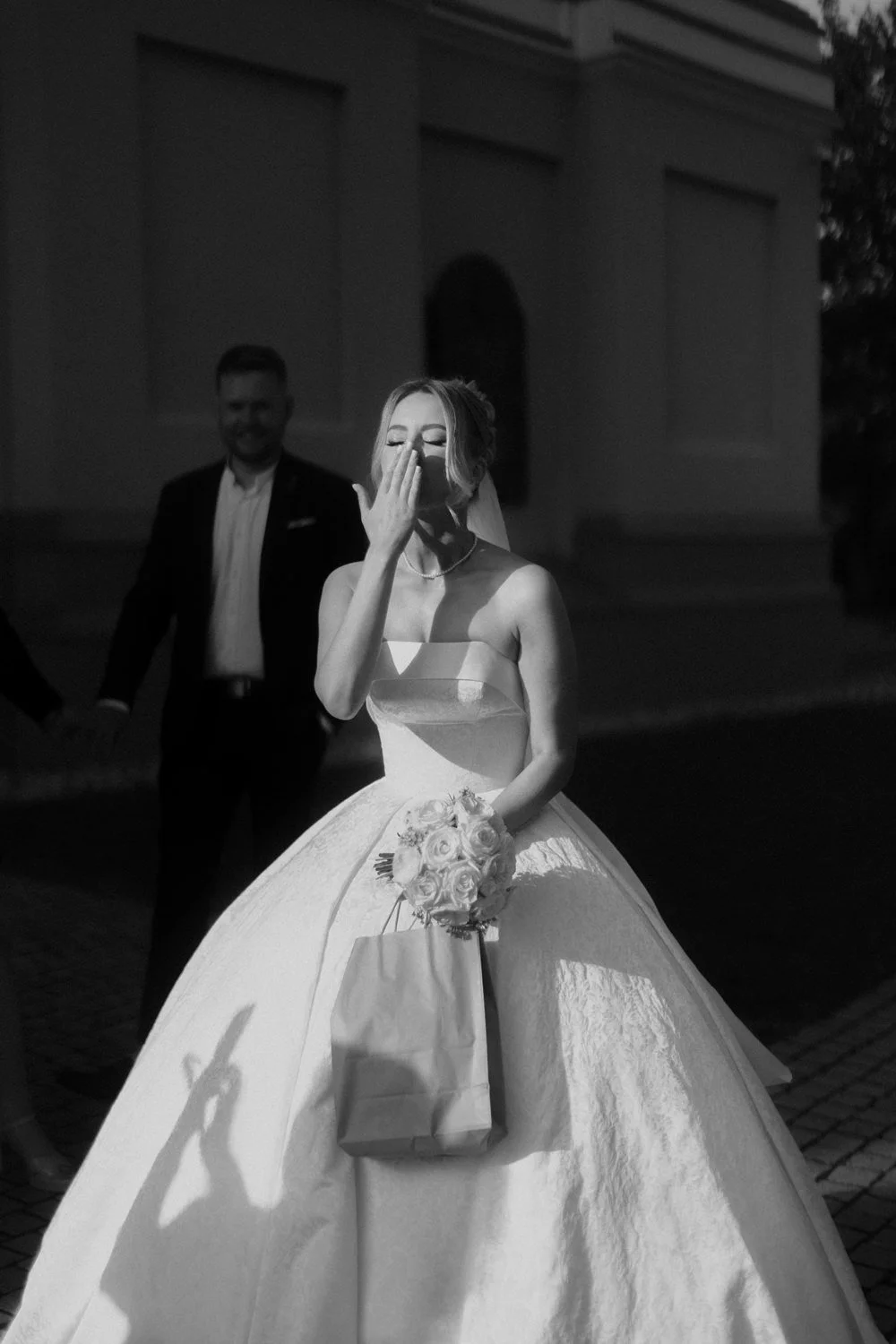 A bride in a wedding gown holding a bouquet of roses after the ceremony, tearing up, with a groom smiling in the background, outside a building.