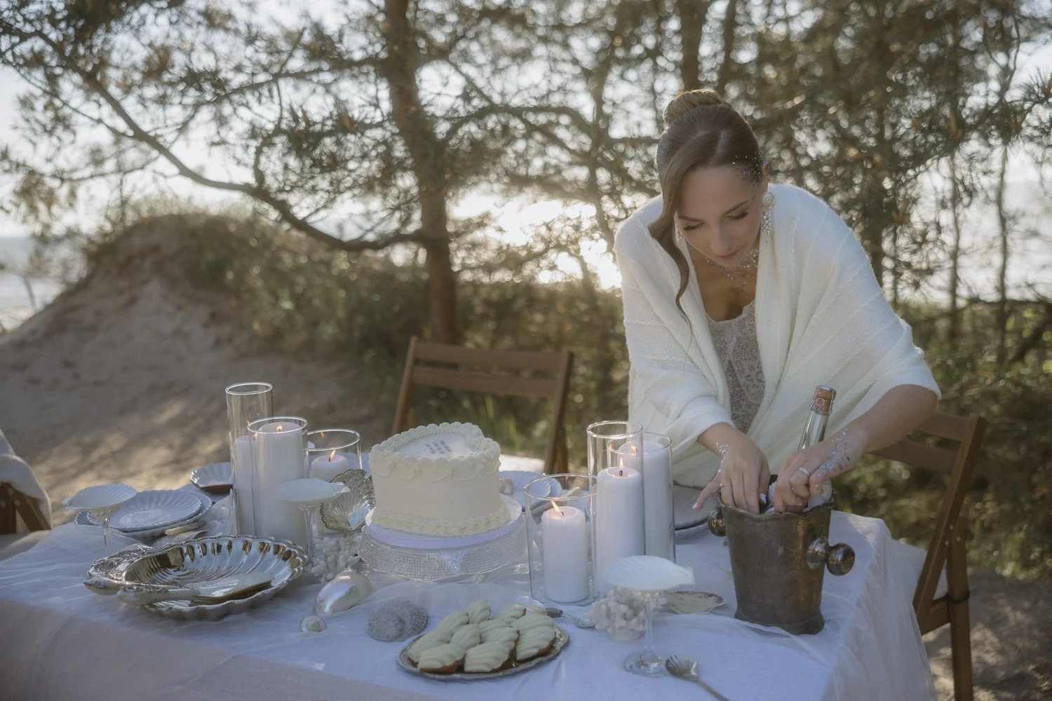 A woman in white attire is preparing candles on a table outdoors, which is decorated with a white cake, candles, and treats. The setting appears to be a beach with trees in the background.