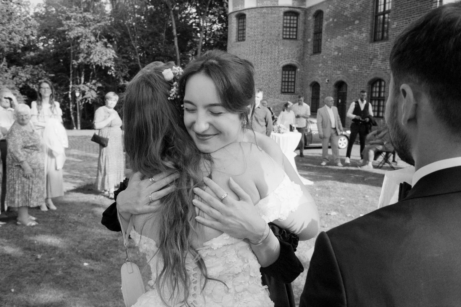 A bride embraces a woman outdoors during a wedding reception, with guests in the background.