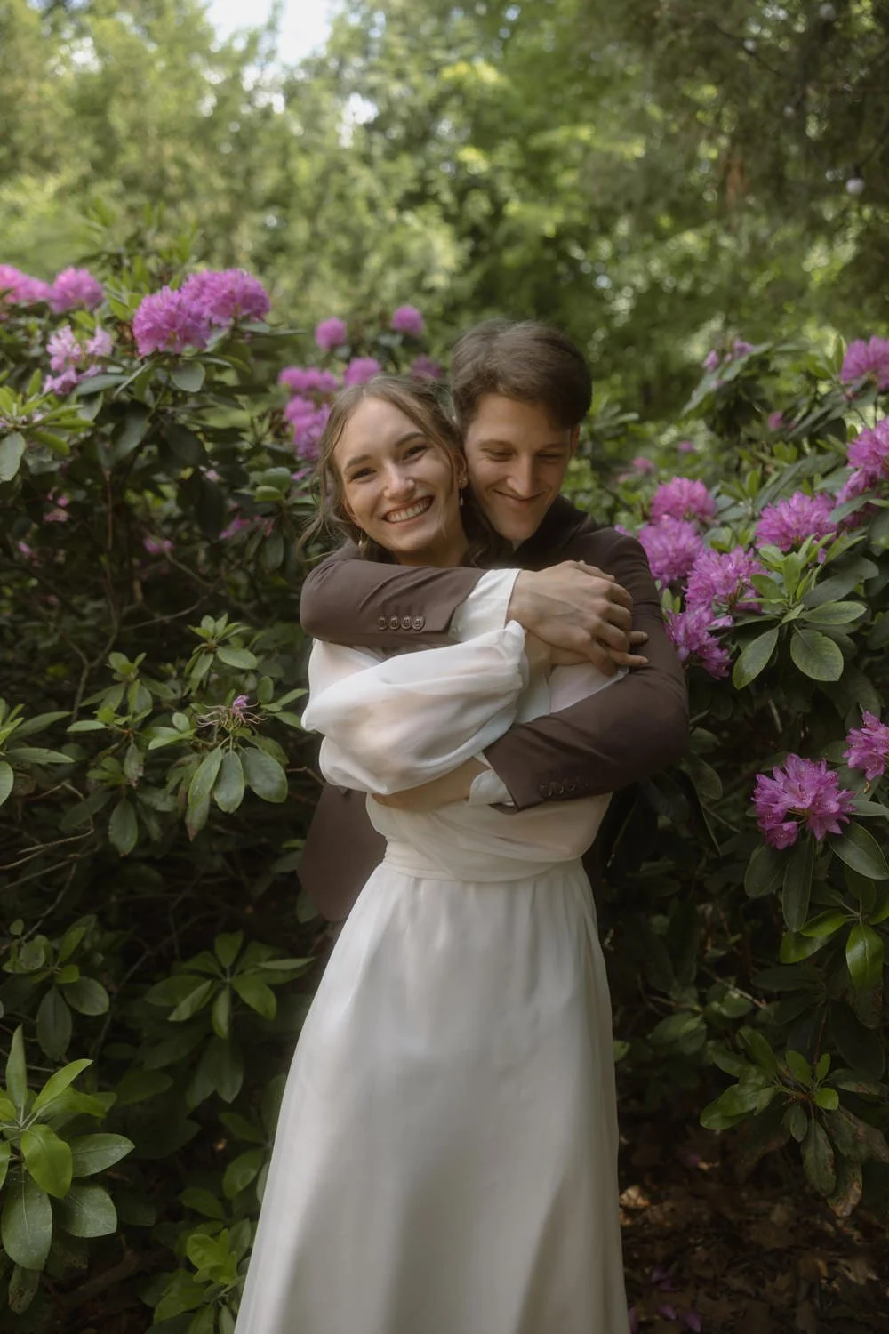 A couple hugging in a garden with pink flowers and green foliage, the woman in a white dress and the man in a brown suit.