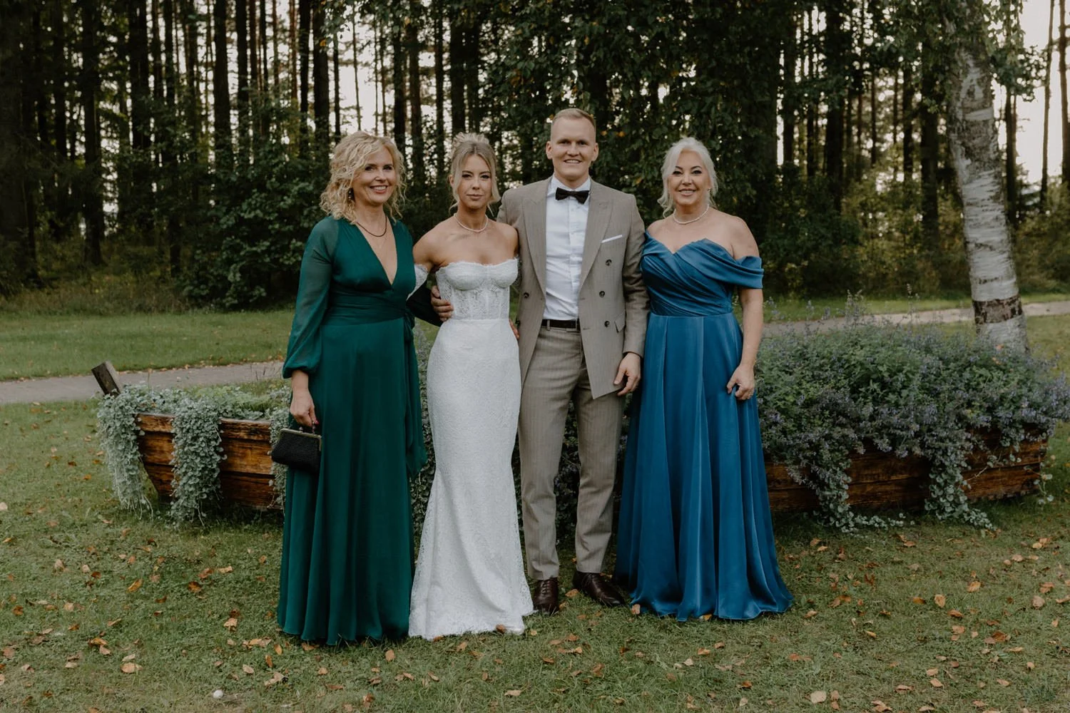 Group of four people dressed in formal attire standing outdoors on grass with trees in the background. One woman in a white wedding dress, the others in teal and blue dresses, and a man in a beige suit and black bow tie.