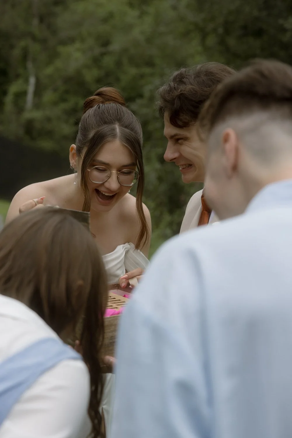 Group of people outdoors, smiling and looking into a box, celebrating a special occasion, with a background of trees and greenery.