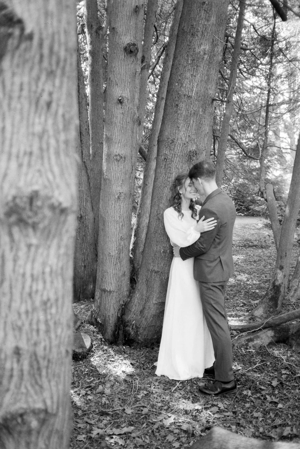 A black and white photo of a couple standing close together in a wooded area, smiling and touching foreheads, with trees surrounding them.